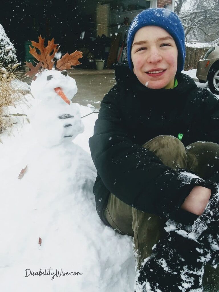 young boy with disabilities sitting next to a small snowman with leaves for hair and carrot nose for the holidays