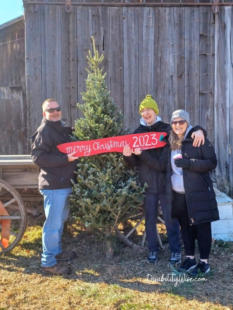 father, son with a disability, and mother at a tree farm holding a holiday sign that says Christmas 2023