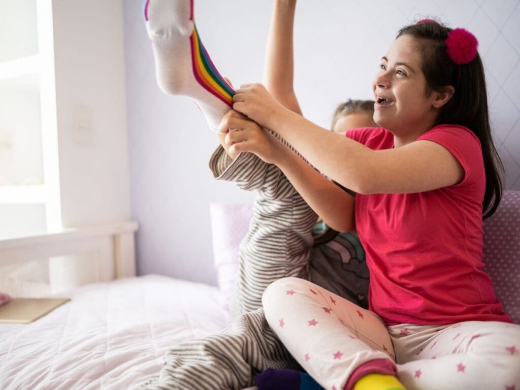 A girl with developmental disabilities helping another girl put on her sock and laughing